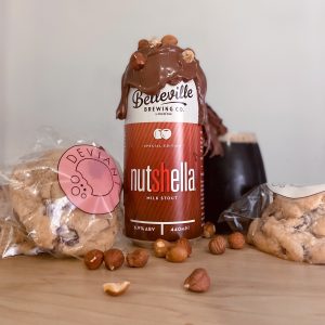 Introducing Nutshella Milk Stout. Photograph of a can of nutshella surrounded by chocolate chip cookies and hazelnuts, with molten chocolate dripping from the top of the can and glass