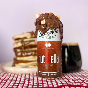 Fluffy American Pancakes Recipe. Photograph of a can of Nutshella covered in chocolate sauce, with a glass of Nutshella stout in the background and a stack of chocolate covered American pancakes on a pink background.