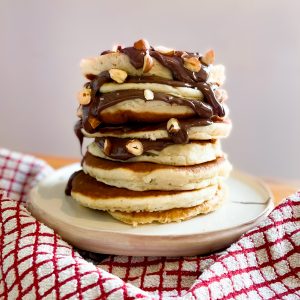 Fluffy American Pancakes Recipe. Photograph of a large stack of American pancakes with chocolate sauce and hazelnuts on top