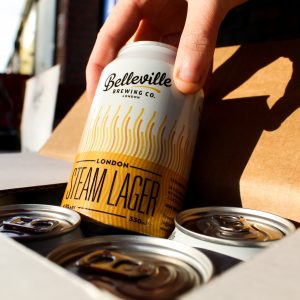 Deep Dive into London Steam Lager. Close up photograph of someone picking a can of London Steam Lager out of a case of beer.